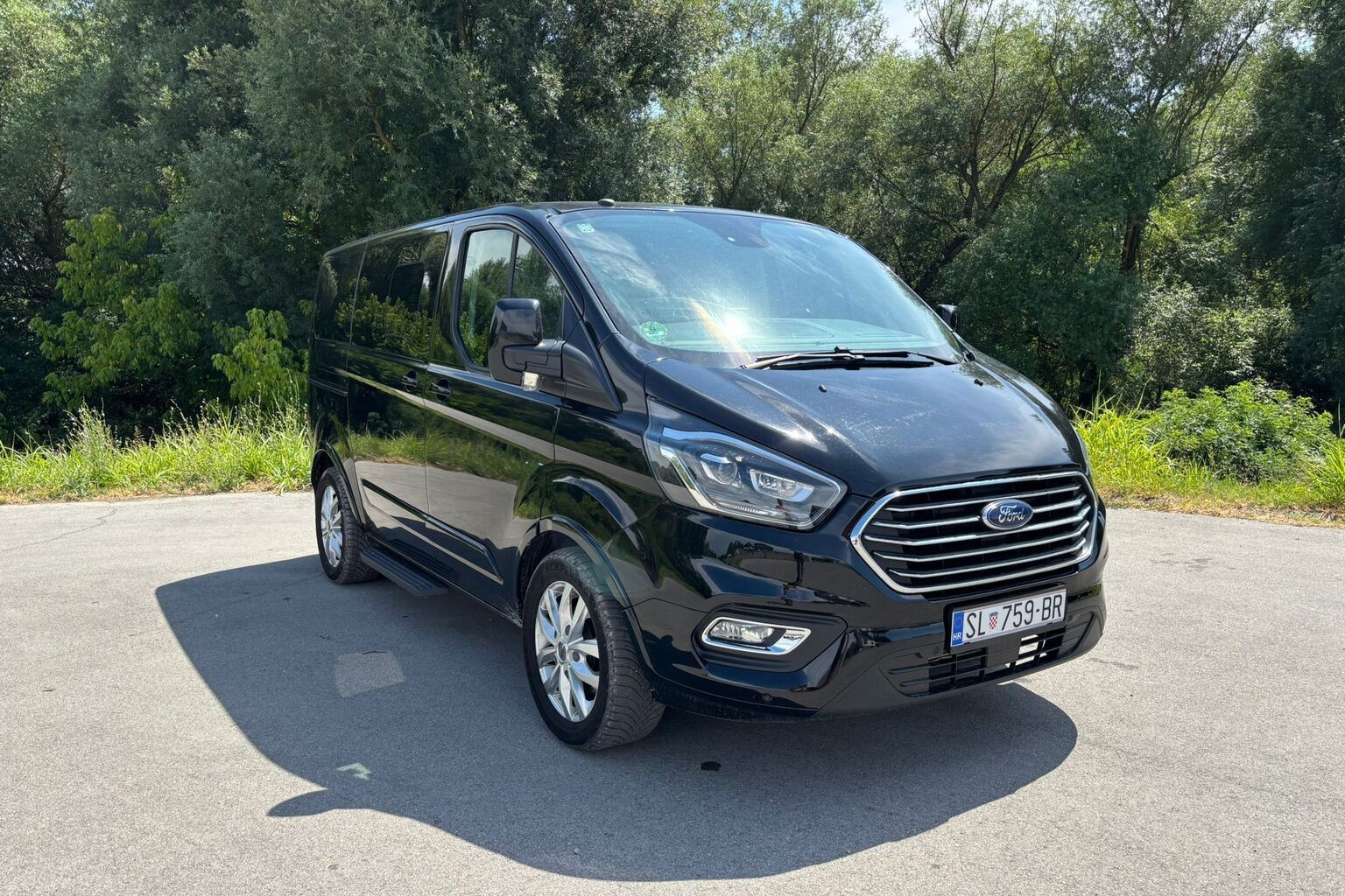Crni kombi Ford Tourneo snimljen sprijeda iz tri četvrtine ispred zelenog drveća