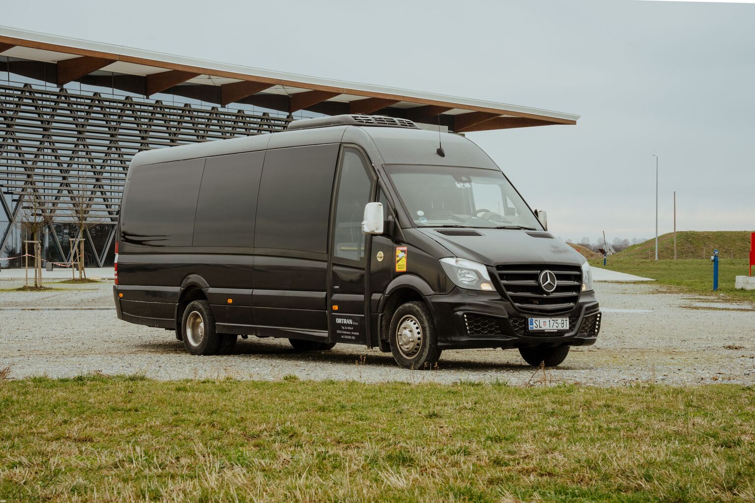 Crni minibus Sprinter snimljen sprijeda iz tri četvrtine na šljunčanom parkiralištu
